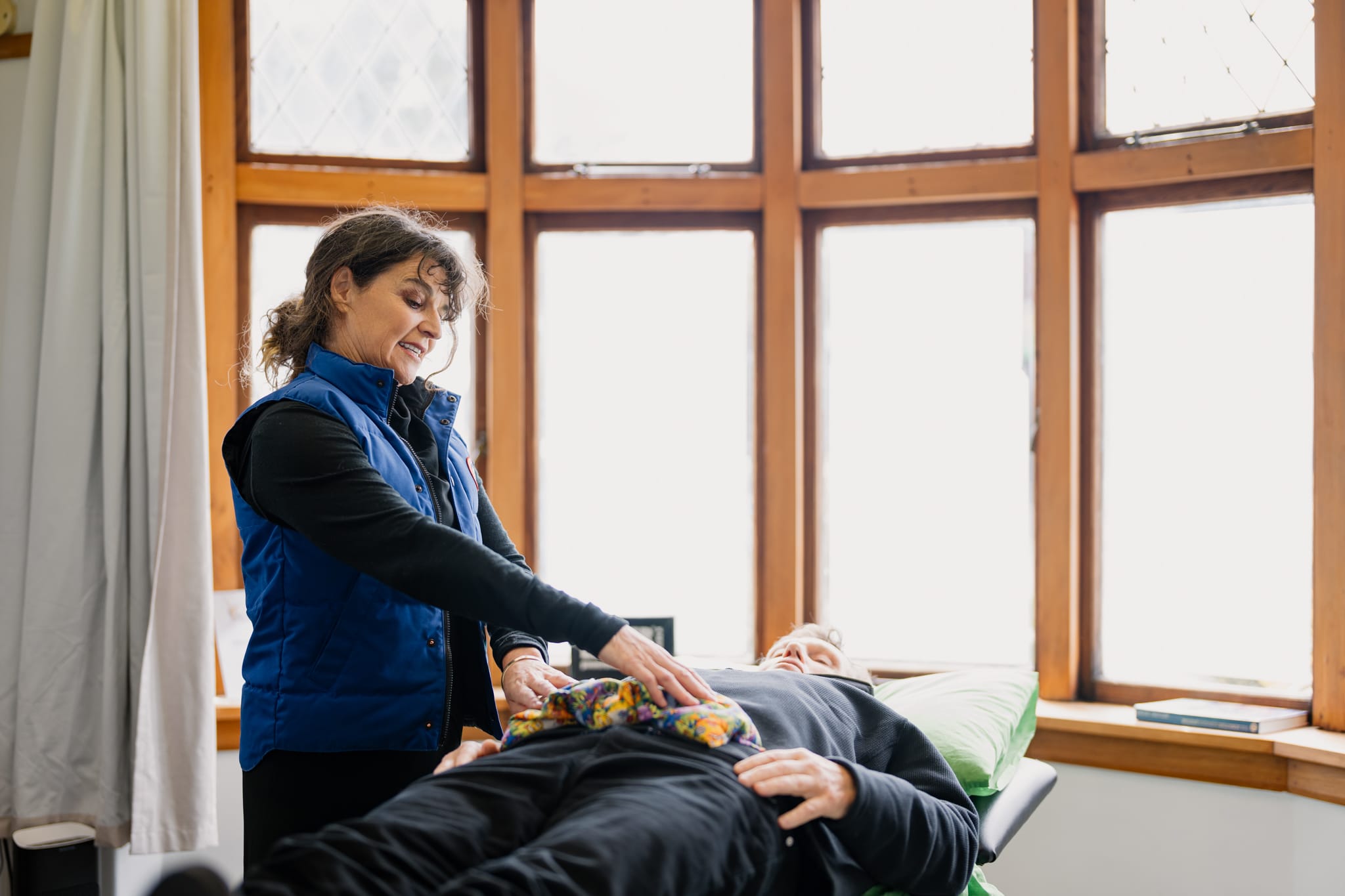 Physiotherapist attending to a patient lying on an examination table near a bright window, placing a wheat bag on the patient's abdomen.